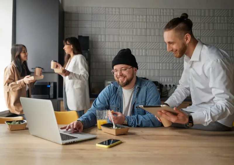 Team Collaborating over Lunch in Bright Kitchen Hub Areas Colleagues enjoying lunch together in a bright kitchen space with laptops, food and drinks business directory